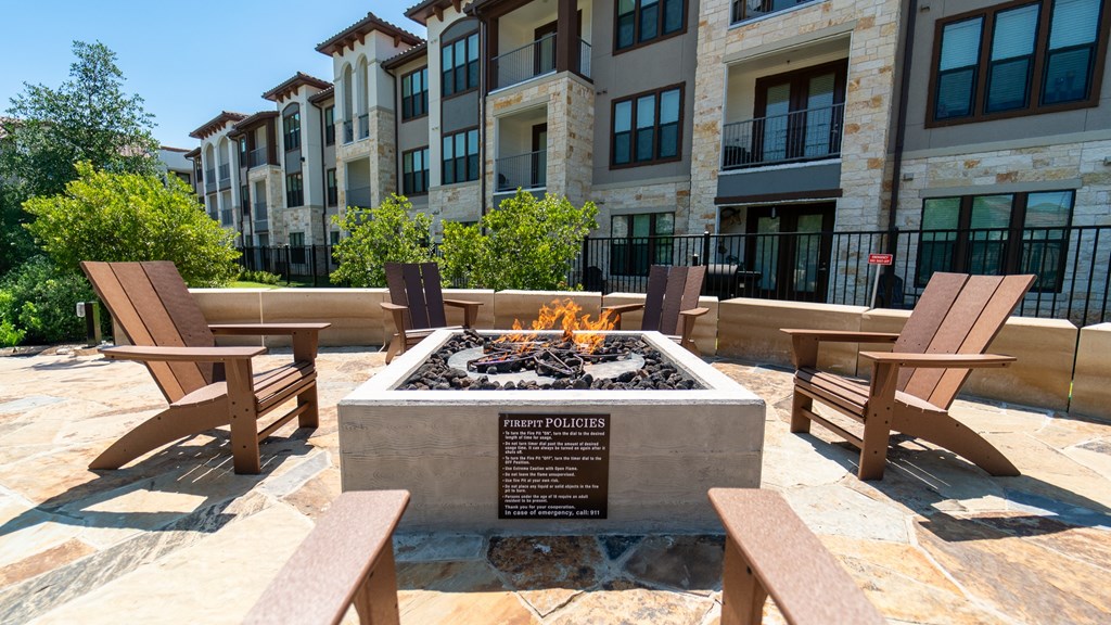 A fire pit is surrounded by wooden chairs in front of a building.