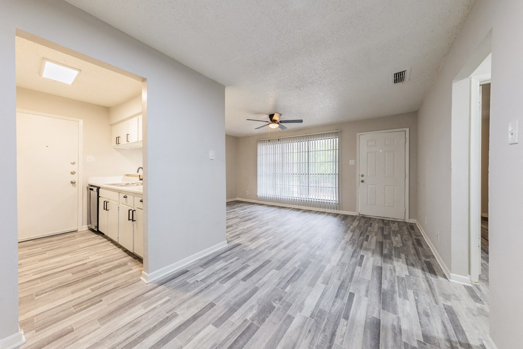an empty living room with a ceiling fan and a kitchen in the background