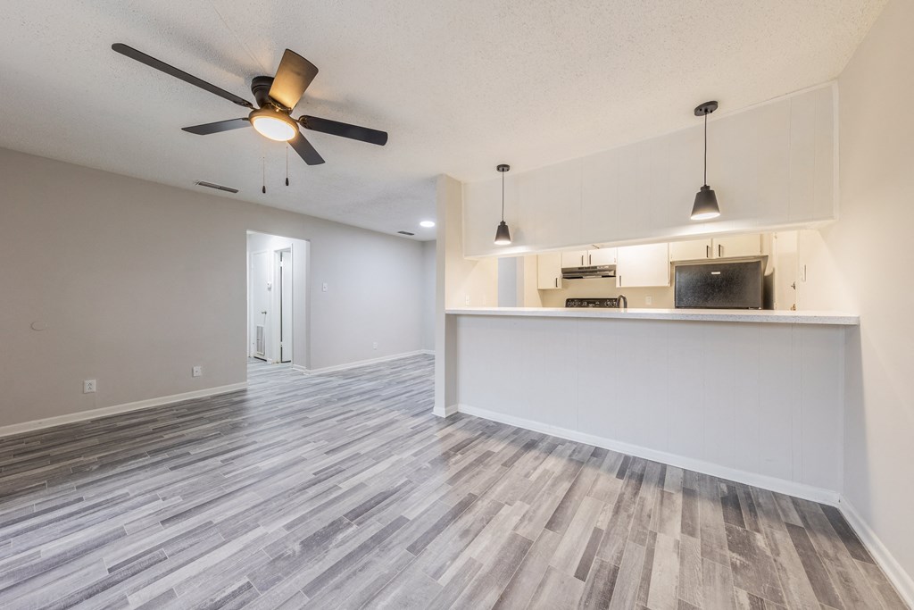 an empty living room with a ceiling fan and a kitchen in the background