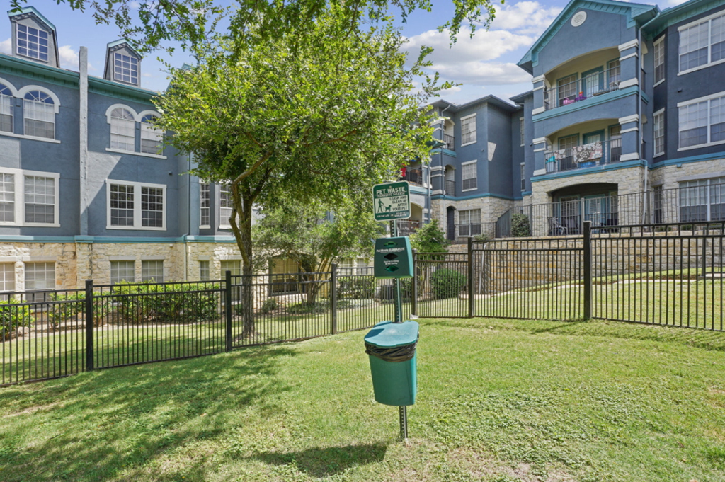 A tree in front of a building with a green trash can in front of it.