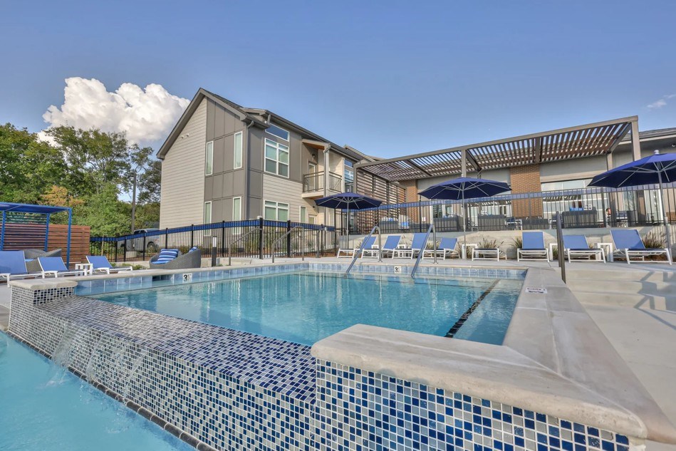A large swimming pool with a blue tiled edge and lounge chairs and umbrellas in front of a building.