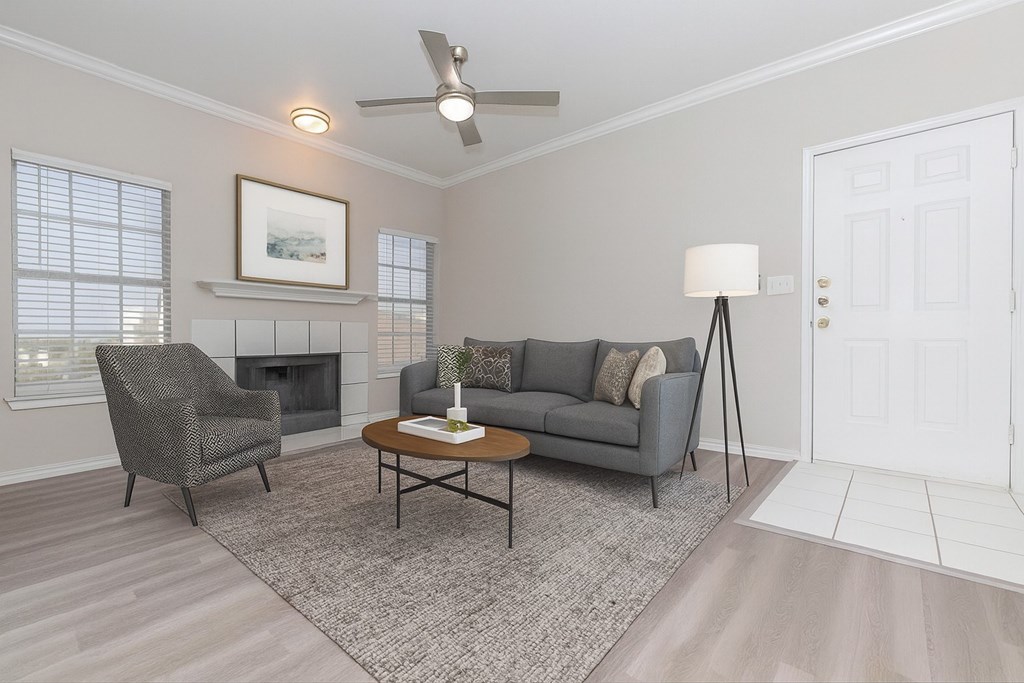 A living room with a grey couch, a grey chair, a grey floor lamp, a grey rug, a grey coffee table, a grey fireplace, a white door, a white wall, and a white ceiling fan.