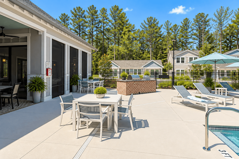 A patio with a table and chairs is surrounded by a pool and trees.