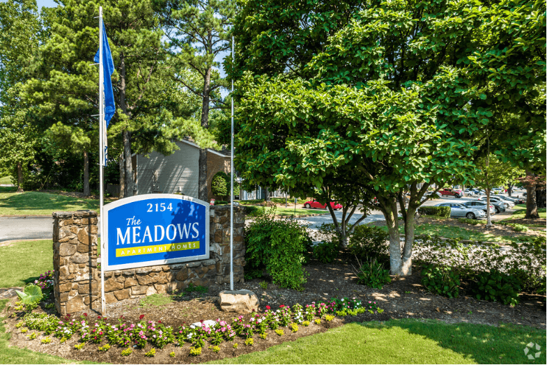 a sign with a blue flag and a stone wall with flowers and trees in the background