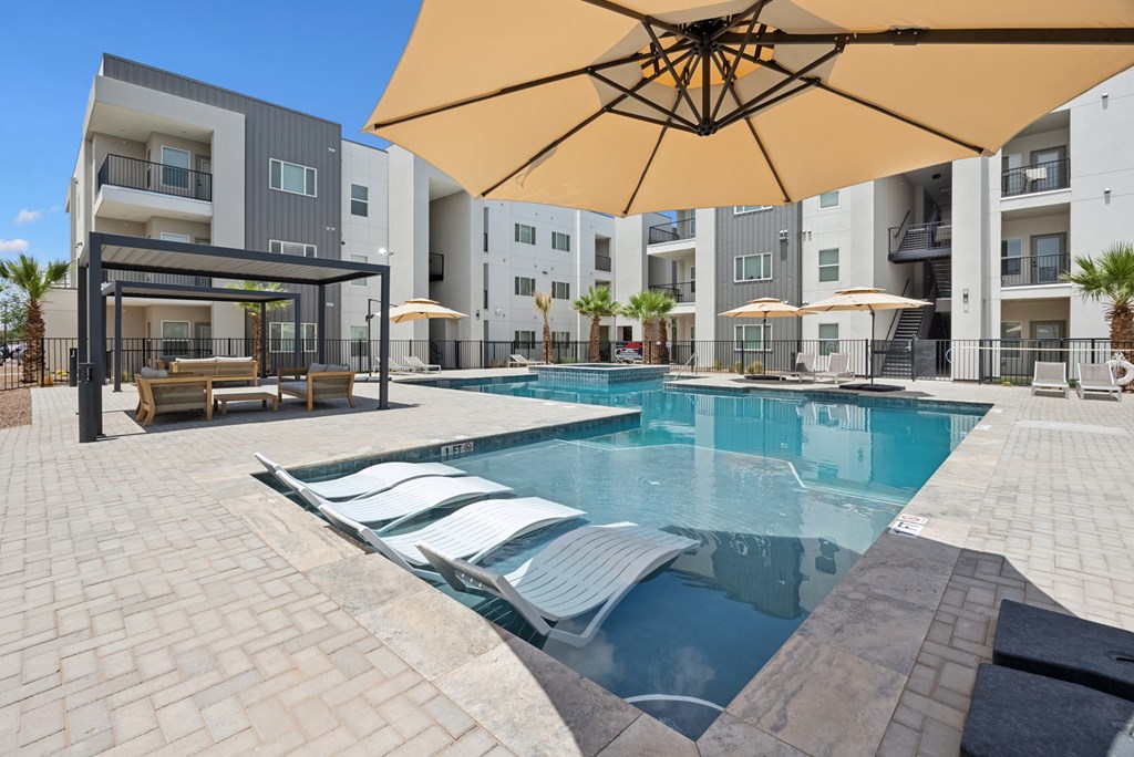 A large umbrella shades a swimming pool area in front of apartment buildings.