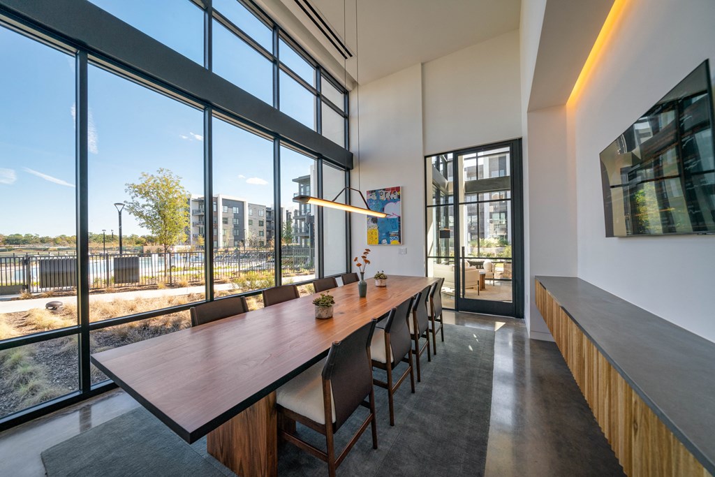 A long wooden table with chairs is in front of a large window with a view of a building and trees.