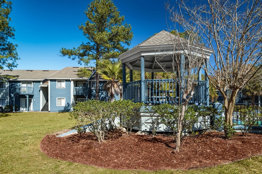 A gazebo is in the middle of a grassy area with a tree and shrubs around it.