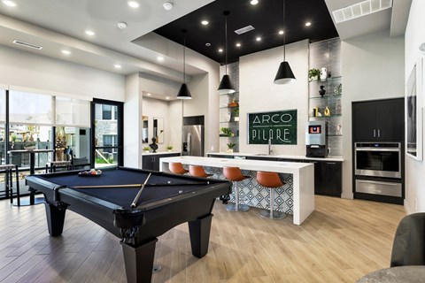 A black pool table is in the middle of a room with a bar and a kitchen in the background.