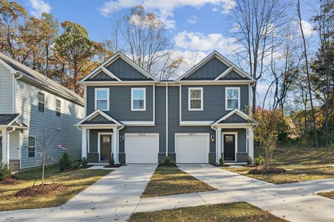 A two-story house with a garage is surrounded by trees and other houses.