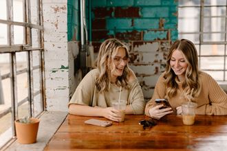 two women sitting at a table drinking coffee and looking at their cell phones