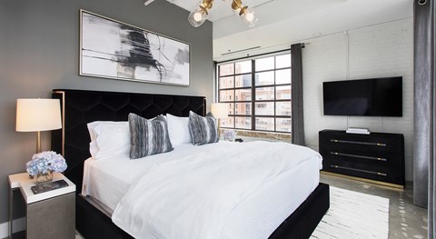 A bedroom with a large bed, a nightstand with a lamp, a side table with a vase of flowers, a TV, and a window with a view of the city.