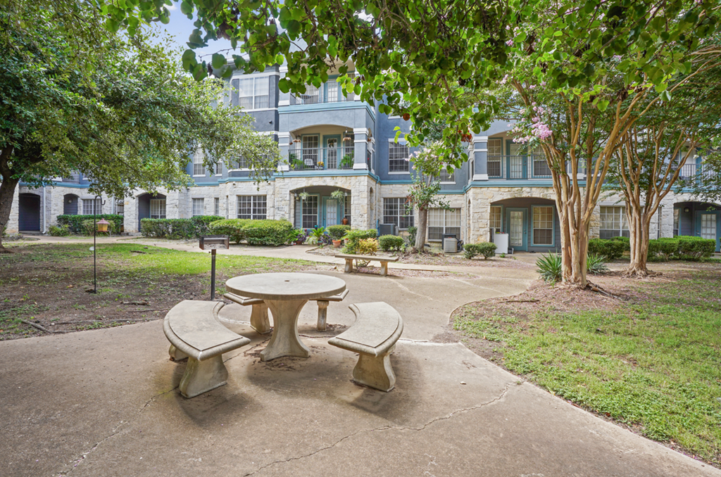 A concrete table and chairs are in the foreground of a courtyard with a building in the background.