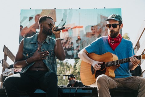 A man in a denim vest is singing into a microphone while another man plays the guitar.
