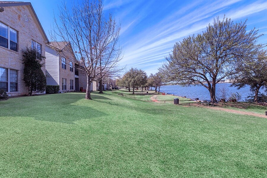A grassy area in front of apartment buildings with a tree and a body of water in the distance.