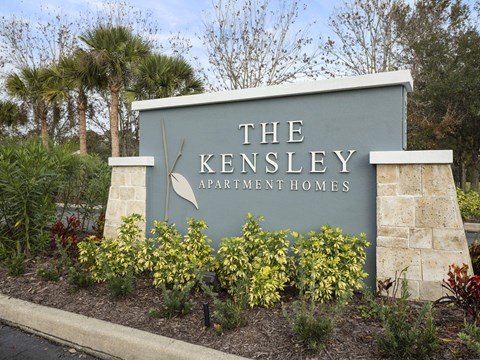 A sign for The Kensley Apartment Homes is surrounded by plants.