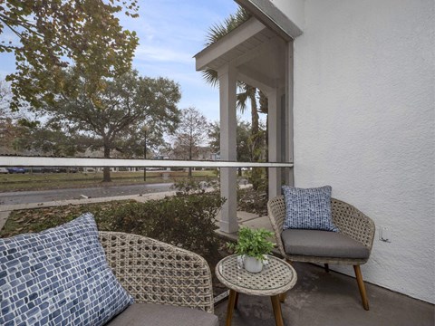 A patio with a wicker chair and a cushioned chair with a pillow and a small table with a plant on it.