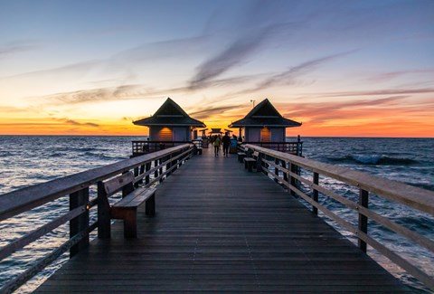 A wooden pier with two pavilions at either end extends into the ocean at sunset.