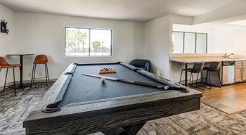 A pool table with a billiard cue and balls is in the foreground of a room with a dining table and chairs in the background.