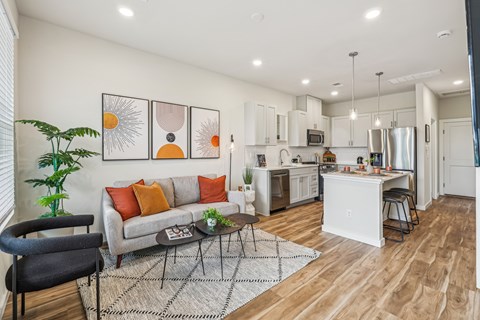 A modern kitchen and living room with a grey couch and orange pillows.