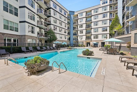 A large swimming pool surrounded by lounge chairs and umbrellas in a courtyard with apartment buildings in the background.