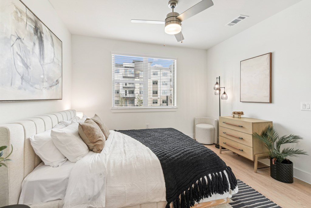 A bedroom with a large bed, a ceiling fan, and a window with a view of a building outside.