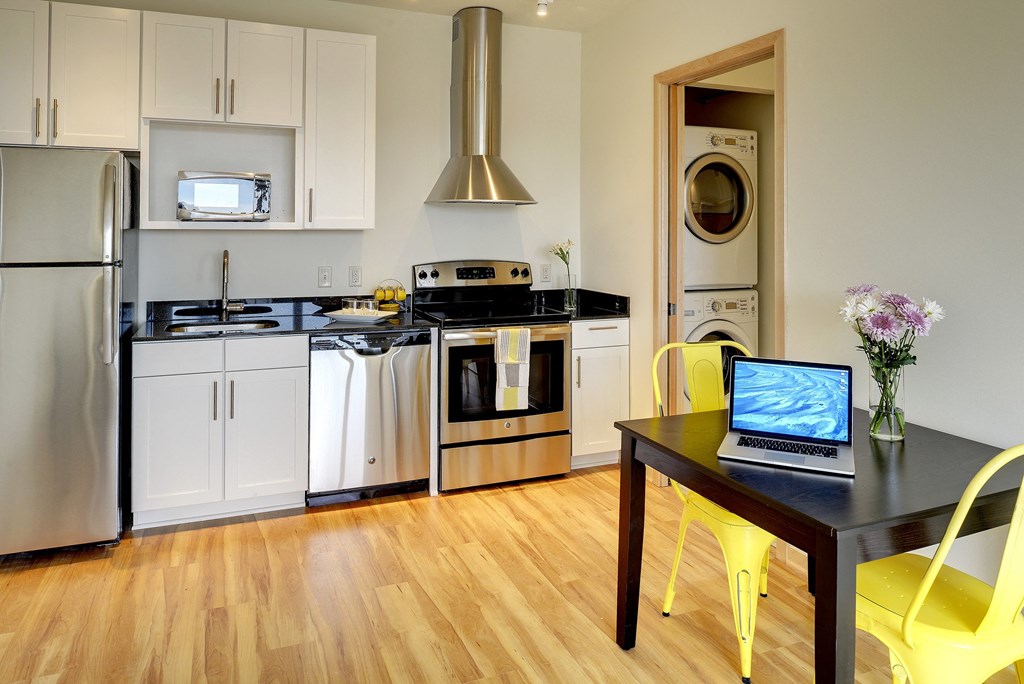 Kitchen with white cabinets, stainless steel appliances and black granite countertops; table with yellow chairs; glimpse into bathroom with washer dryer stack