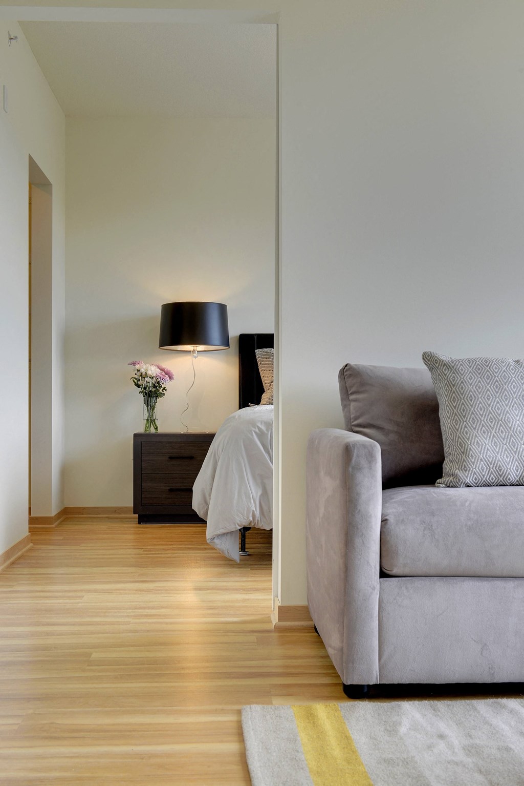 Flat J living area with grey couch and glimpse into bedroom behind couch with black nightstand and lit lamp