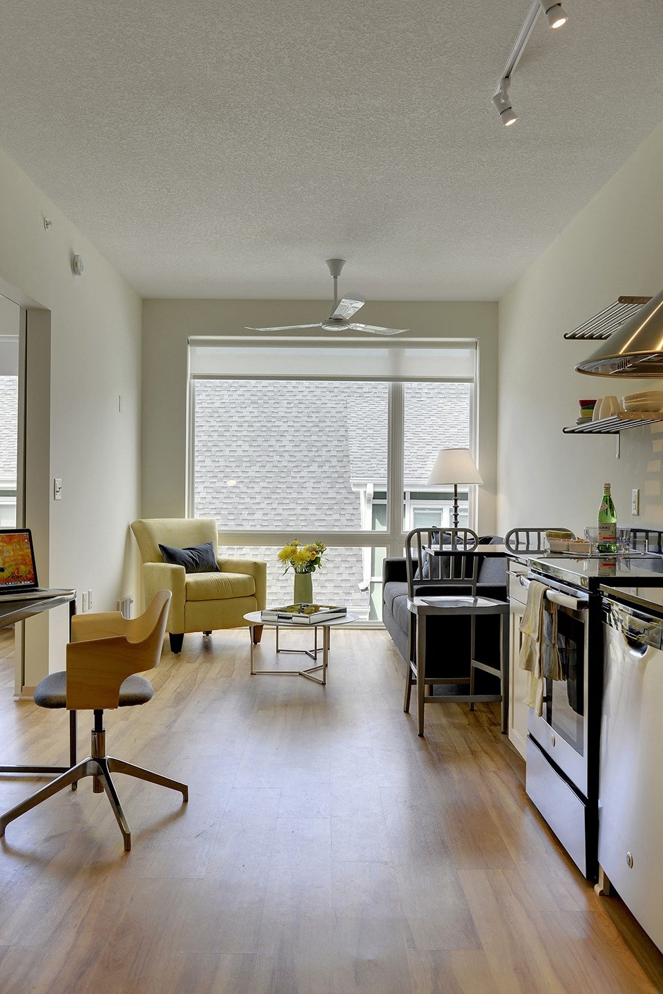 Living space with desk nook, yellow accent chair, grey couch and glimpse of kitchen in the Flat E floor plan at Coze Flats