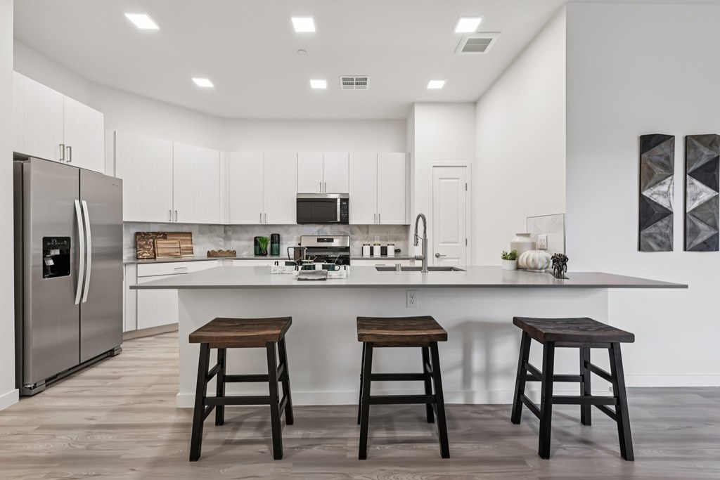 A kitchen with white cabinets and a white countertop with a refrigerator on the left and two stools in front of the counter.