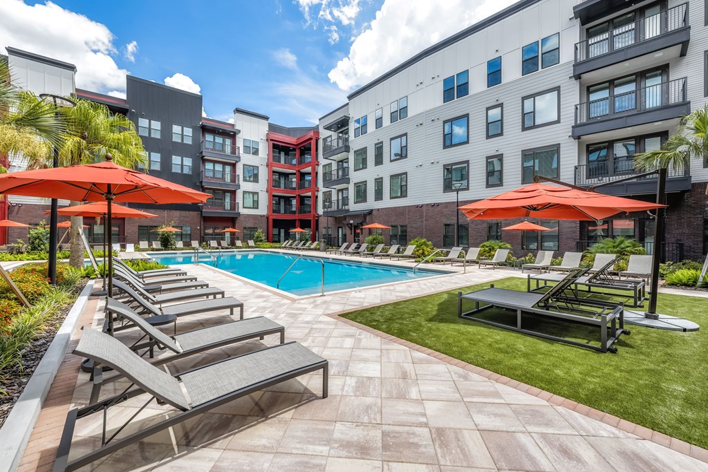 A pool surrounded by chairs and umbrellas with apartment buildings in the background.