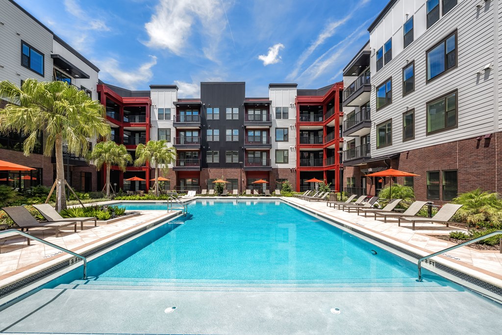 A swimming pool in front of a multi-story building with lounge chairs around it.