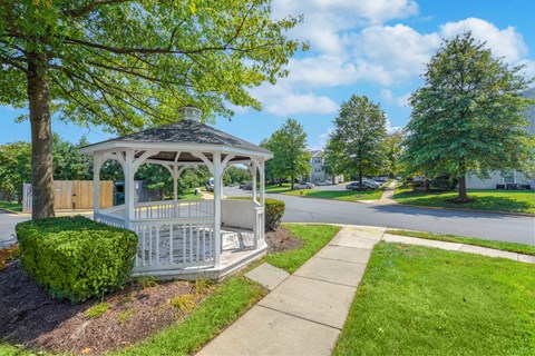 A white gazebo with a black roof is surrounded by greenery and a sidewalk.