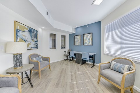 A living room with a grey sofa, a grey armchair, a grey chair, a round table, a lamp, a painting, a cabinet, a desk, and a window with blinds.