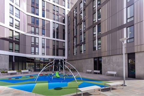 A playground with a blue and green surface is surrounded by modern buildings.