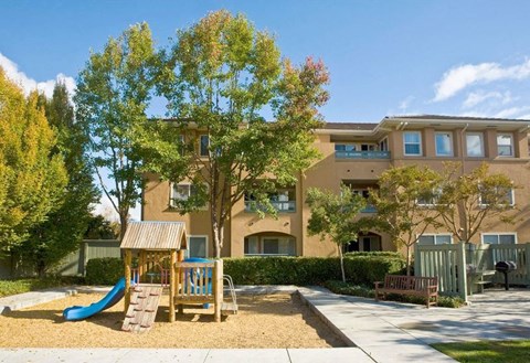 A playground with a slide and a wooden structure is in front of a building.