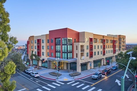 A multi-story building with a red top floor is situated on a street corner with cars parked in front.