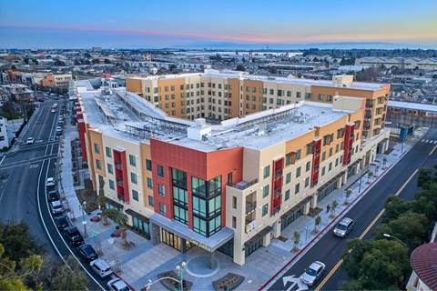 A large, modern building with a curved facade is surrounded by a parking lot and a street with cars.
