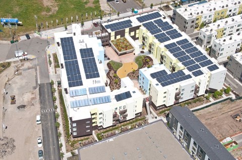 A building with solar panels on the roof is surrounded by other buildings.