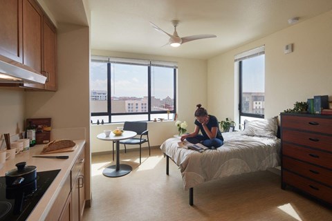 A woman is sitting on a bed in a room with a kitchen area.