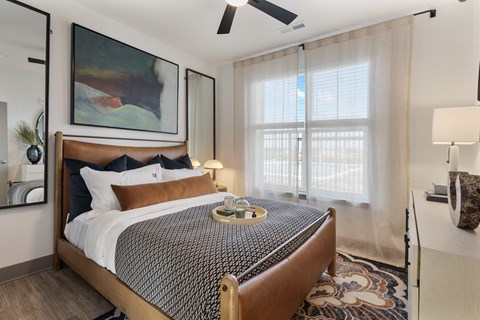 Model apartment bedroom at Montage West Lafayette featuring a modern bed frame, layered bedding, ceiling fan, and large window with natural light.