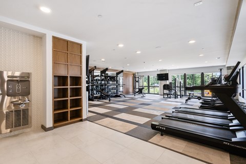 Modern apartment gym fully equipped with cardio machines, weight training equipment, a flat-screen TV, individual storage cubbies, and a convenient resident hydration station.