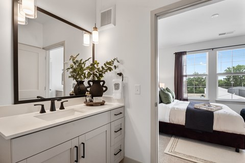 A view from a modern bathroom vanity featuring a light wood cabinet and quartz countertop, looking through an open doorway into a bright, carpeted bedroom with large windows.