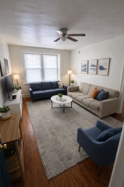 A living room with a grey rug, a blue armchair, a grey sofa, a white coffee table, a wooden cabinet, a television, and a ceiling fan.