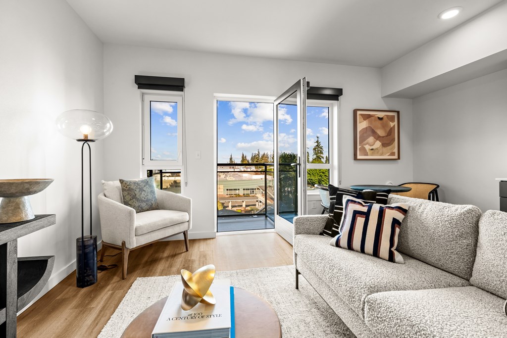 A living room with a grey couch, a white chair, a table with a book and a gold object on it, and a view of the outside through the windows at Two 10 by Vintage, Shoreline, WA