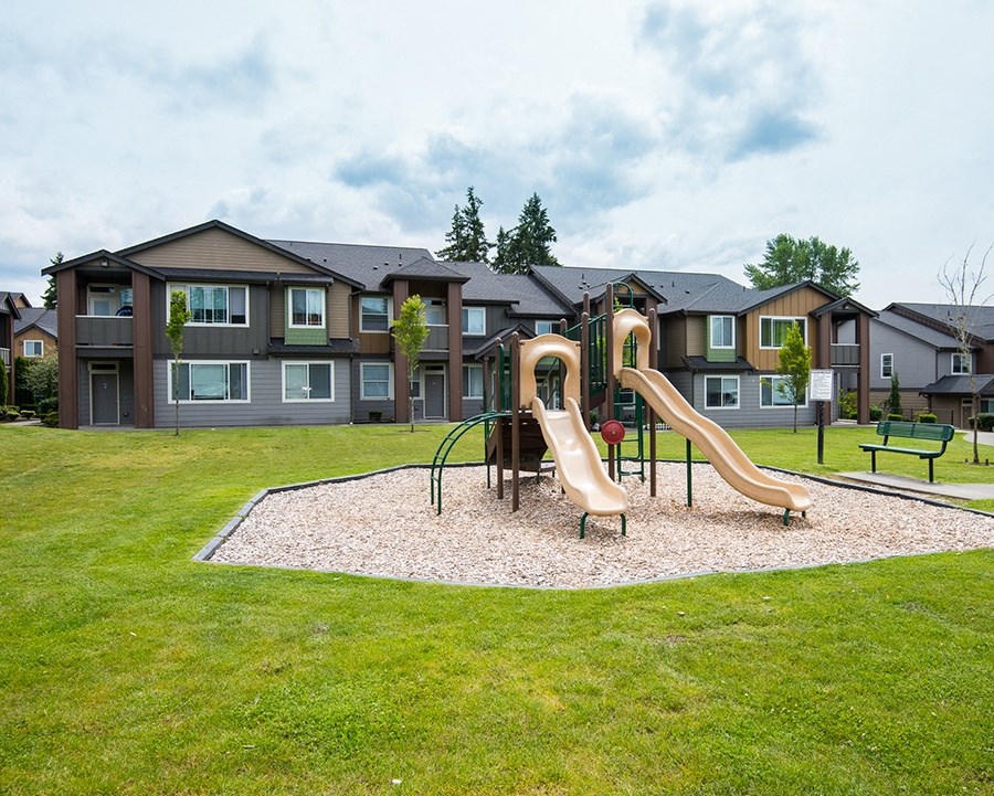 South Hill Puyallup Apartments. - Bella on Canyon - Playground Surrounded by Grass with Climbing Structure that Includes Two Slides and a Rounded Climbing Ladder