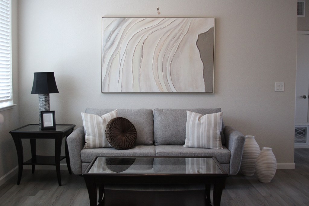A living room with a grey couch, a table with a lamp and a picture frame on it, and a large painting on the wall.