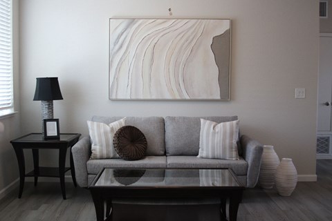 A living room with a grey couch, a table with a lamp and a picture frame on it, and a large painting on the wall.