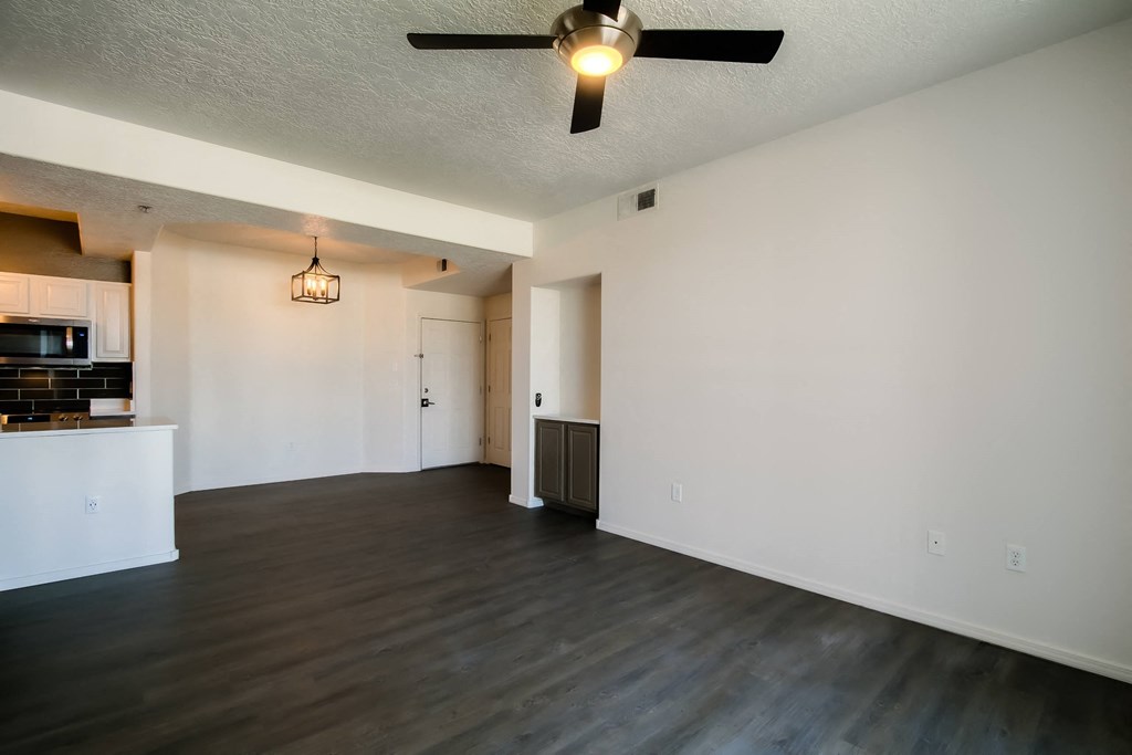 an empty living room with a ceiling fan and a kitchen in the background