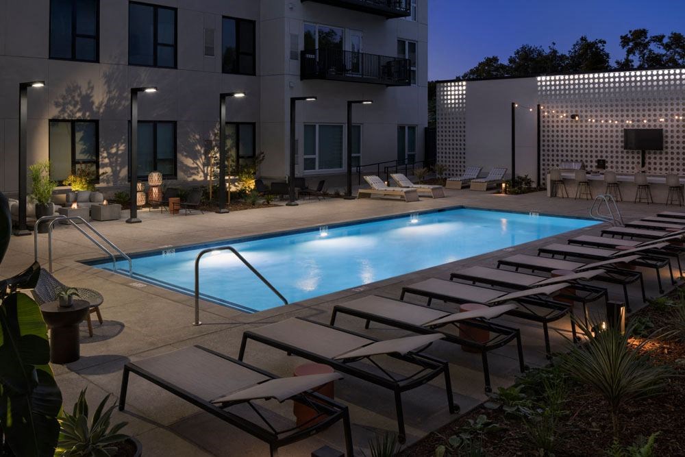 Apartments Near Midtown Sacramento CA - The Mansion - Glowing Blue Pool at Night with Poolside Lounge Chairs, Plants, String Lights, TV, and Grill Station