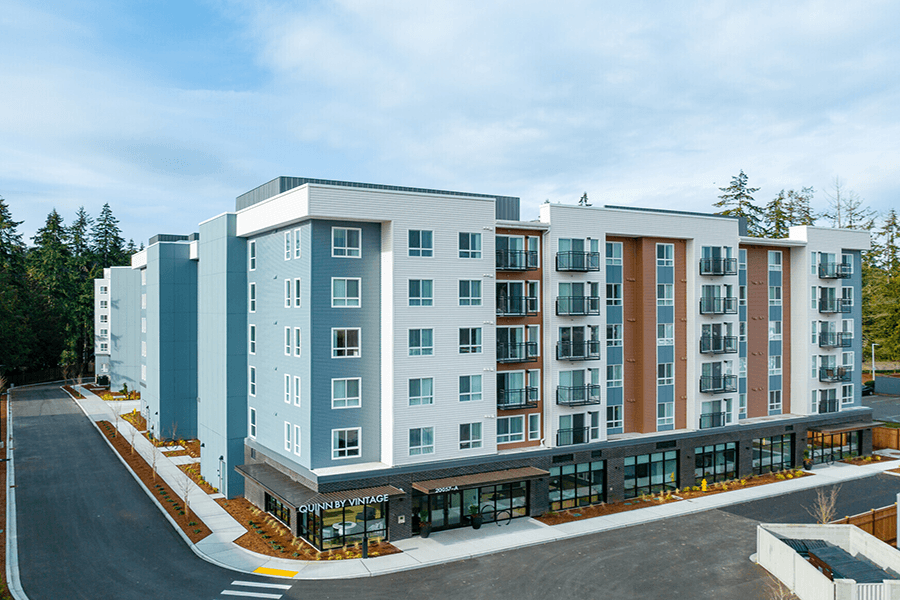 Shoreline Apartments for Rent - Quinn by Vintage - Exterior or Apartment Complex with Parking Lot Access, Tall Trees, and Blue Skies